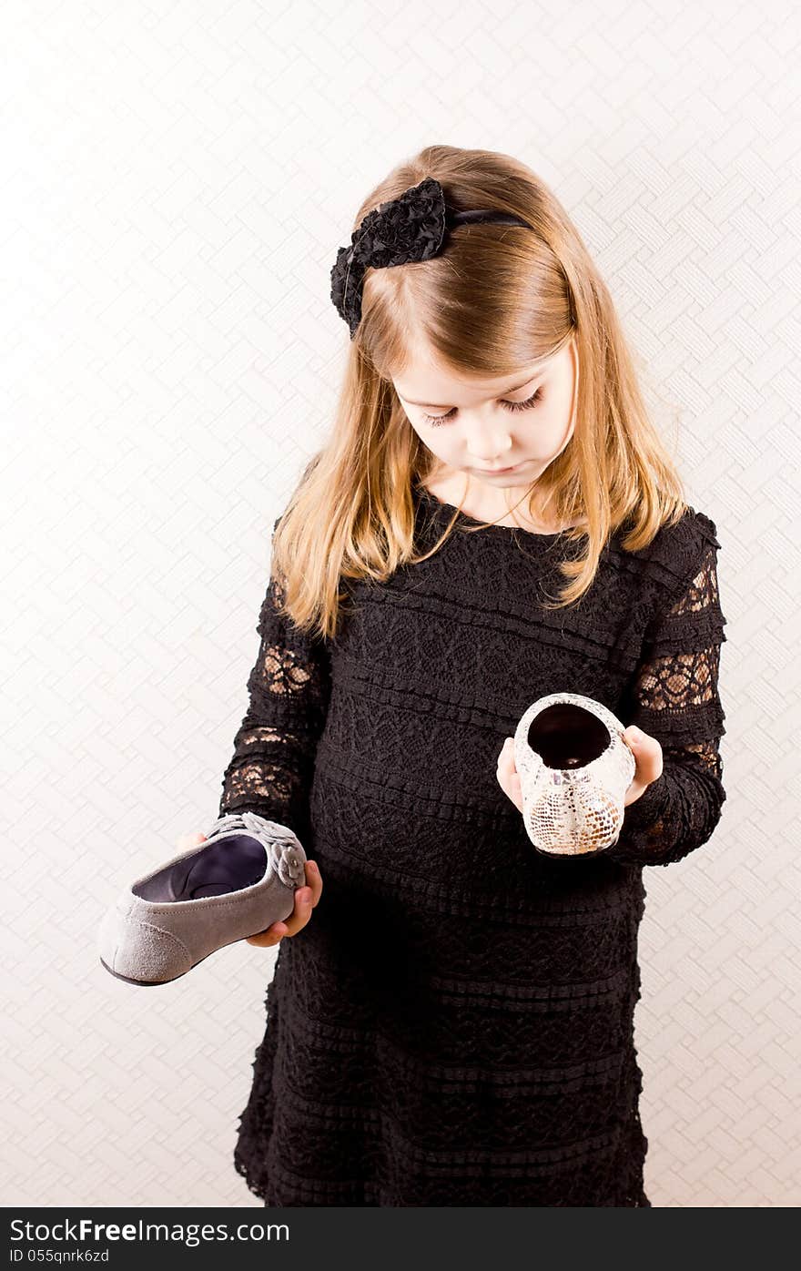 Little blonde girl standing with a different shoe in each hand making a choice of footwear to wear with her stylish black outfit isolated against a neutral background. Little blonde girl standing with a different shoe in each hand making a choice of footwear to wear with her stylish black outfit isolated against a neutral background.