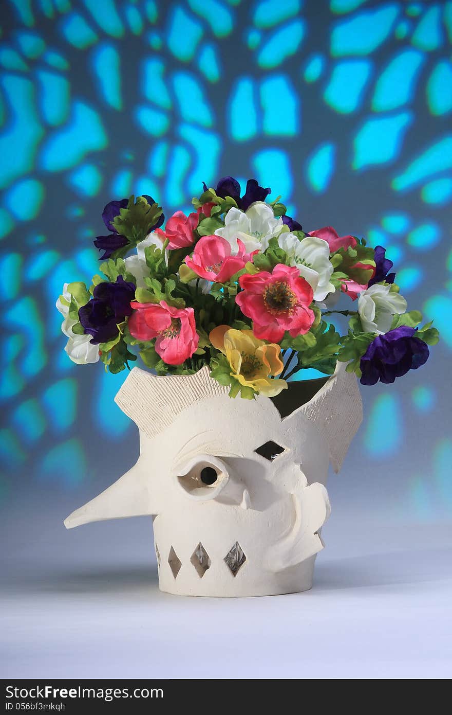 A vase of unglazed ceramic, representing a head with a long nose. Inside the head a bunch of artificial flowers in various colors. Behing there is a light blue background with phantasy structure. A vase of unglazed ceramic, representing a head with a long nose. Inside the head a bunch of artificial flowers in various colors. Behing there is a light blue background with phantasy structure.