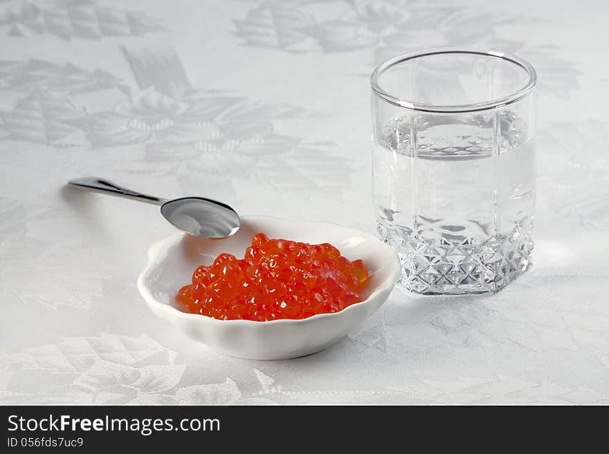 A small glass of vodka, salmon caviar in a plate for caviar and small spoon. A small glass of vodka, salmon caviar in a plate for caviar and small spoon