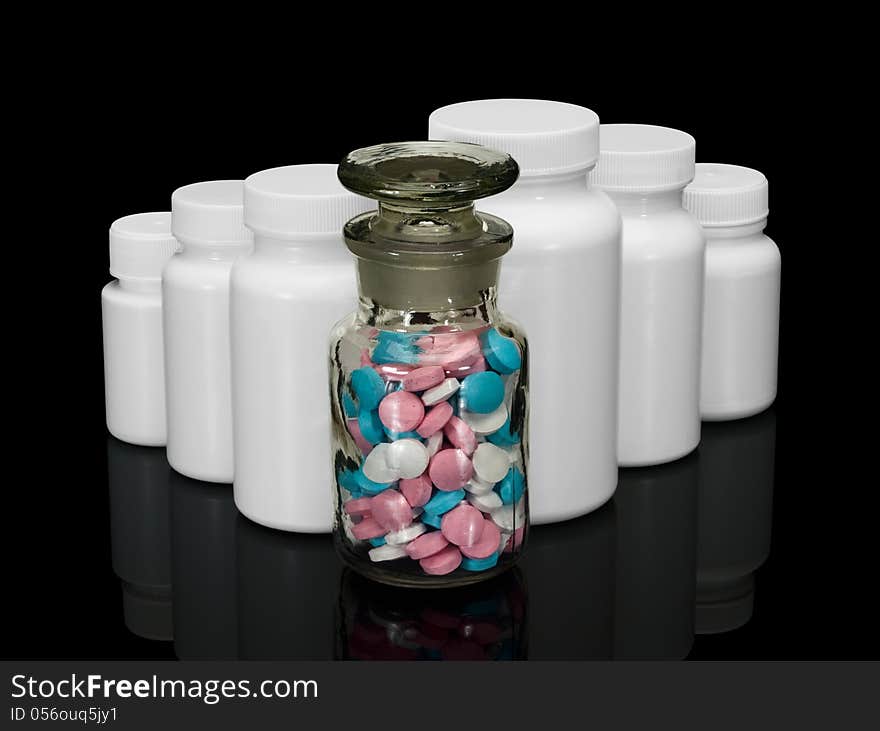 Glass bottle with multi-colored pills and row of white small bottles for pills on a black background. Glass bottle with multi-colored pills and row of white small bottles for pills on a black background.