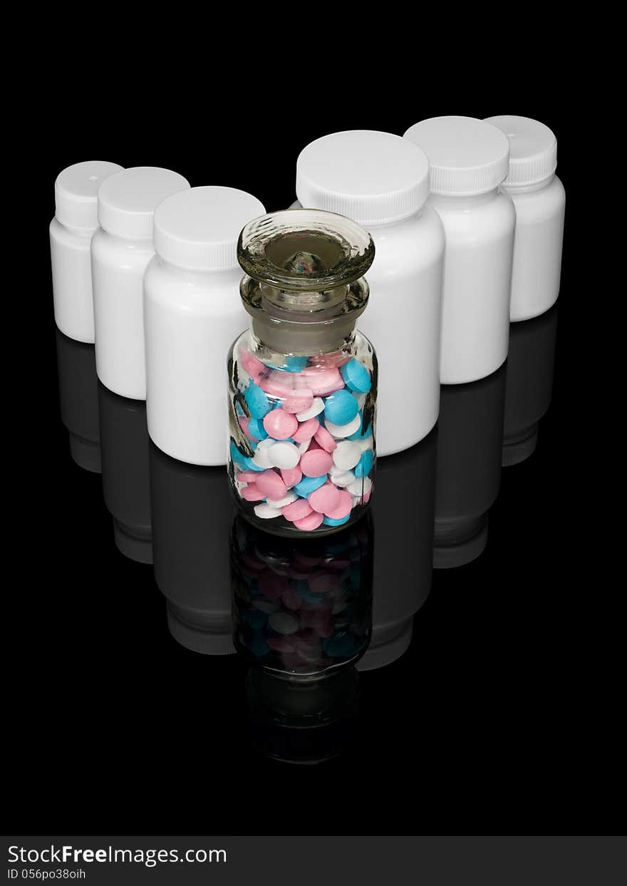 Glass bottle with multi-colored pills and row of white small bottles for pills on a black background. Glass bottle with multi-colored pills and row of white small bottles for pills on a black background.