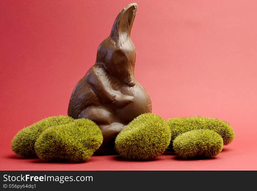 Australian alternative to the Easter bunny rabbit, a chocolate Bilby holding an egg on green moss rocks against a red background.