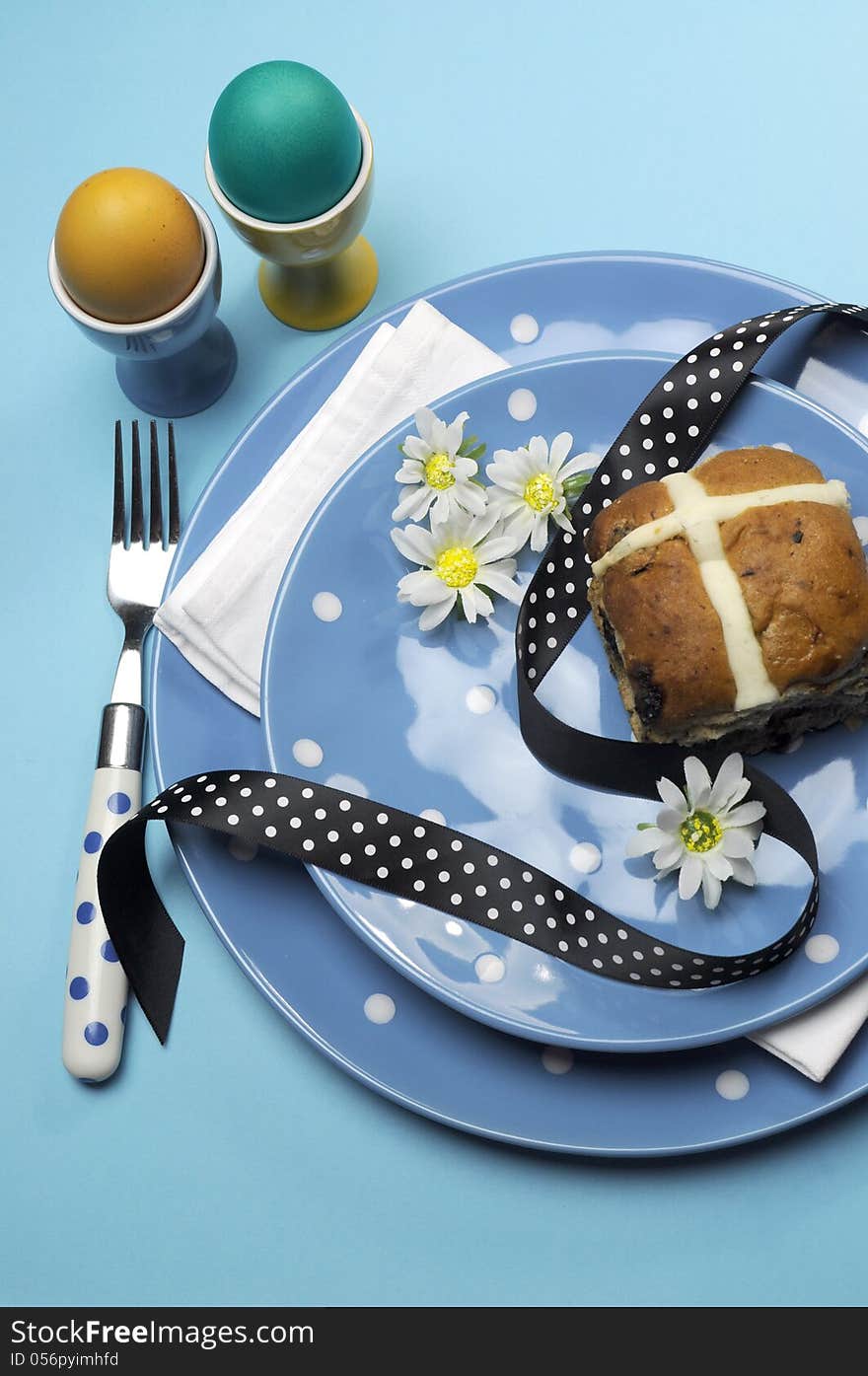 Happy Easter dinner table setting with blue polka dot plates, and decorations against a blue background. Vertical with Easter Hot Cross Bun and color boiled eggs. Happy Easter dinner table setting with blue polka dot plates, and decorations against a blue background. Vertical with Easter Hot Cross Bun and color boiled eggs.