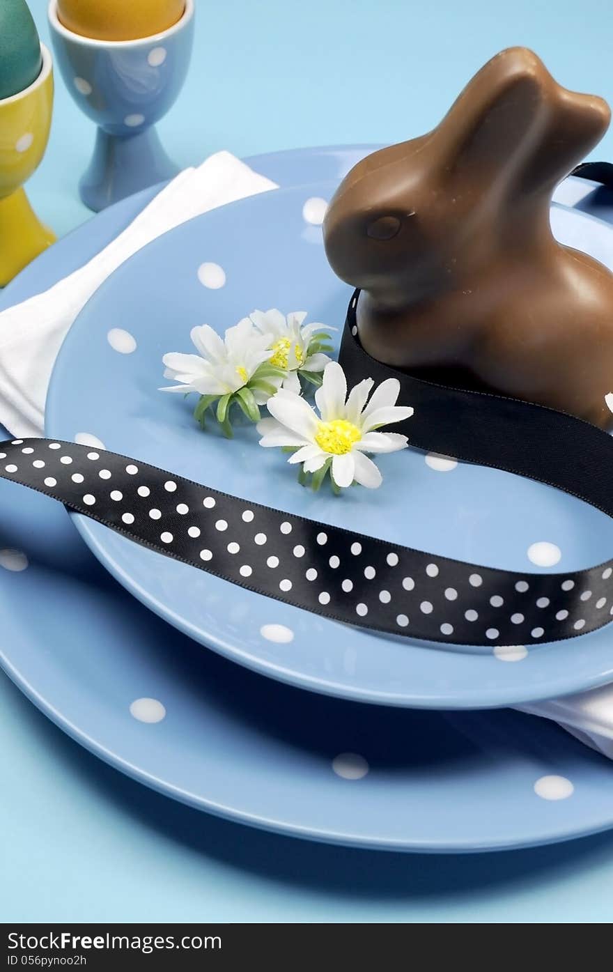 Happy Easter dinner table setting with blue polka dot plates, and decorations against a blue background. Vertical close-up with chocolate Easter bunny rabbit. Happy Easter dinner table setting with blue polka dot plates, and decorations against a blue background. Vertical close-up with chocolate Easter bunny rabbit.