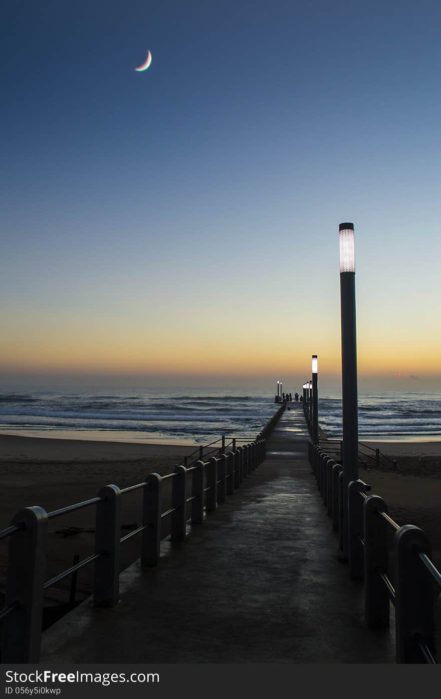 An image of a crescent moon hanging low in the sky during the early morning light over a beach with a pier still light up by light poles. An image of a crescent moon hanging low in the sky during the early morning light over a beach with a pier still light up by light poles.