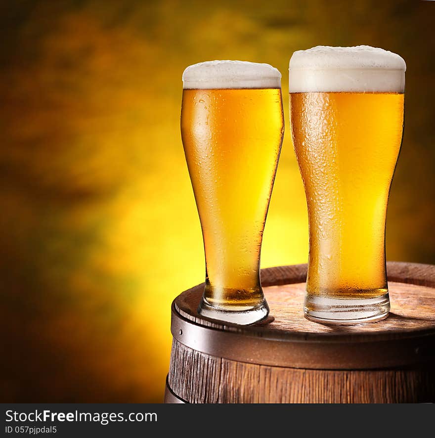 Two glasses of beer on a wooden barrel. Dark yellow background with a gradient.