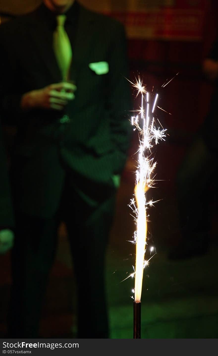 Birthday party. Focus on the sparkler in the foreground. In the background a man wear suits and hold a a glass of champagne.