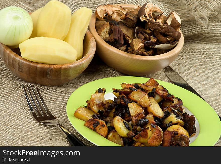 Arrangement of Green Plate with Roasted Potato, Two Wood Bowls with Raw Potato, Onion and Slices of Mushrooms and Fork with Knife closeup on Sackcloth background