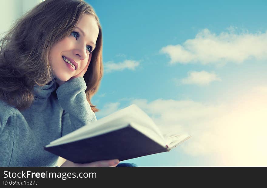 Beautiful young woman reading a book while sitting at a window in the sky and sunset