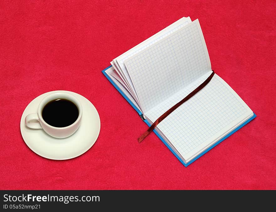 The cup of coffee and open notebook on a red blanket. The cup of coffee and open notebook on a red blanket