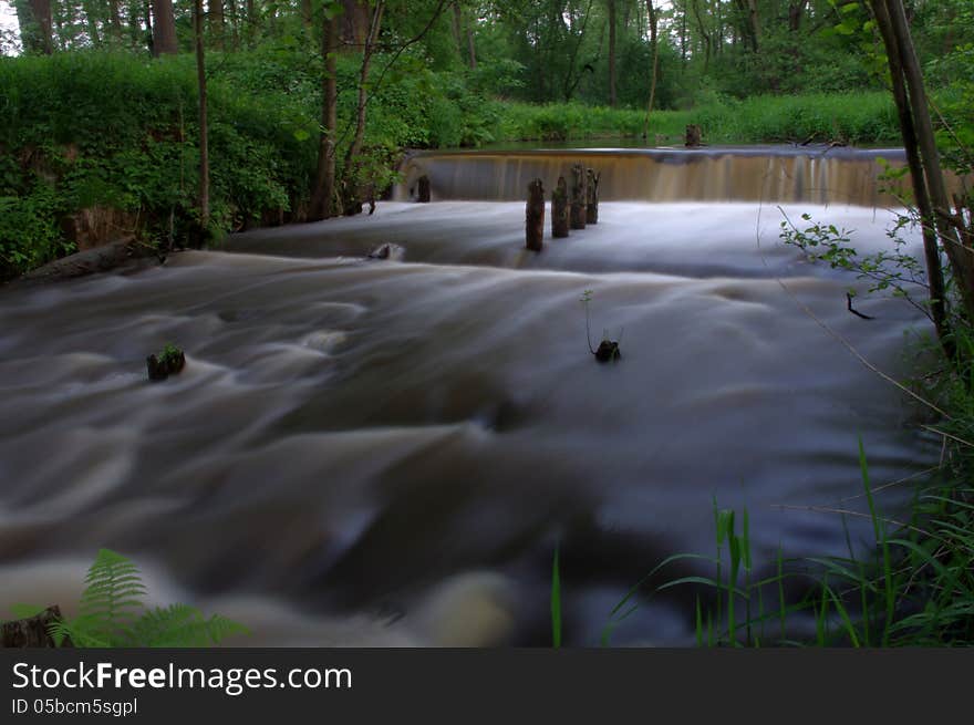 The photo shows a small waterfall on a small river. It is located in the forest, surrounded by greenery. The photo shows a small waterfall on a small river. It is located in the forest, surrounded by greenery.