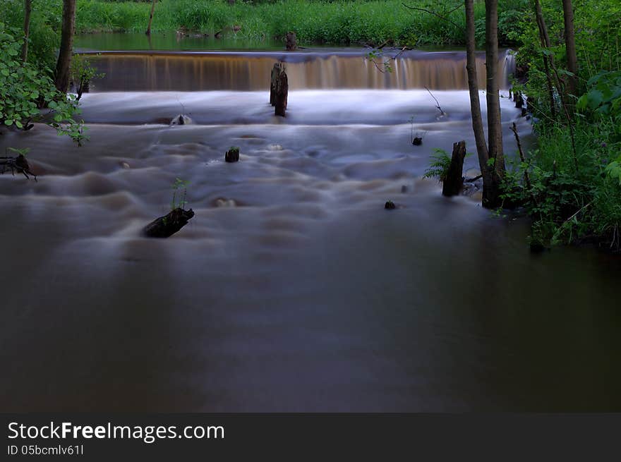 The photo shows a small waterfall on a small river. It is located in the forest, surrounded by greenery. The photo shows a small waterfall on a small river. It is located in the forest, surrounded by greenery.