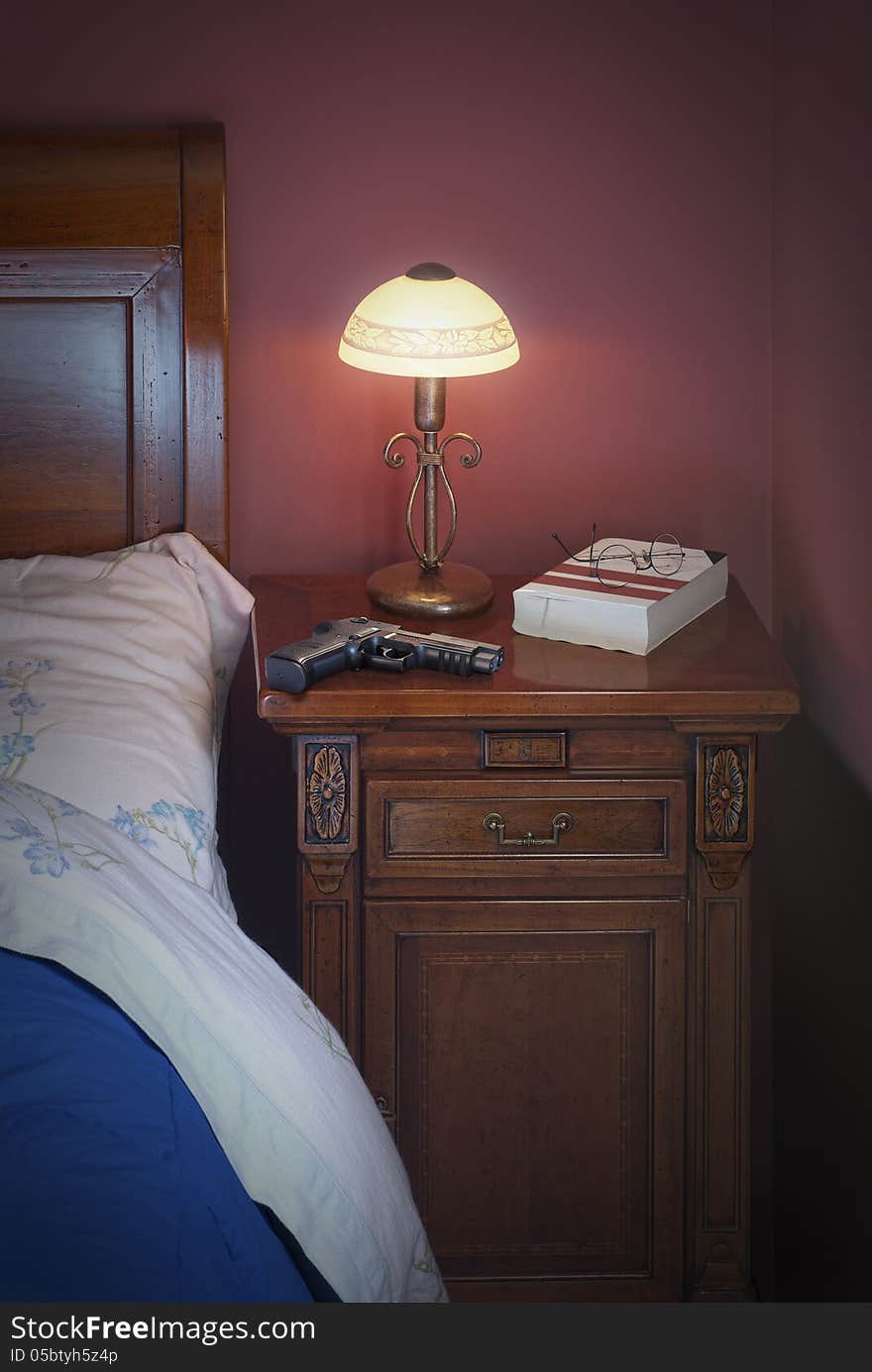 Glock on night table.With a night lamp, book and glasses beside a blu bed. This is ready to read a thriller book maybe and he's afraid to be scared