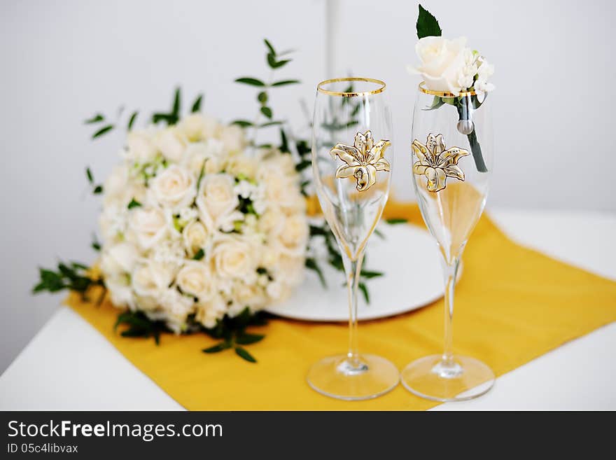 Two decorated wedding glasses with a groom boutonniere and the bride's bouquet in the background. Two decorated wedding glasses with a groom boutonniere and the bride's bouquet in the background