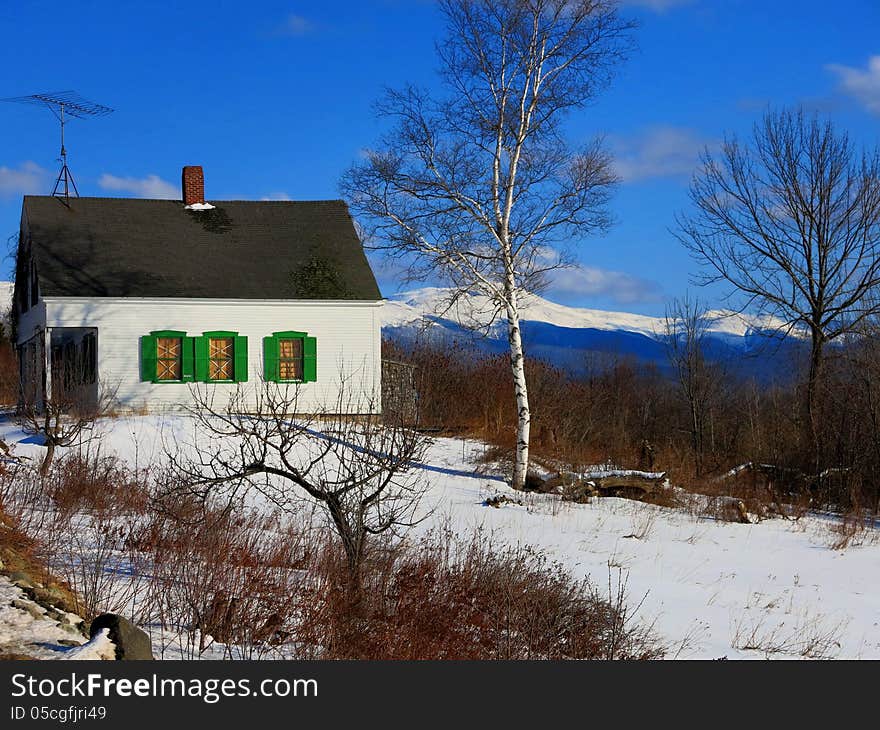 Abandoned house with green shutters in snow with blue sky and birch trees and White Mountains in background. Abandoned house with green shutters in snow with blue sky and birch trees and White Mountains in background