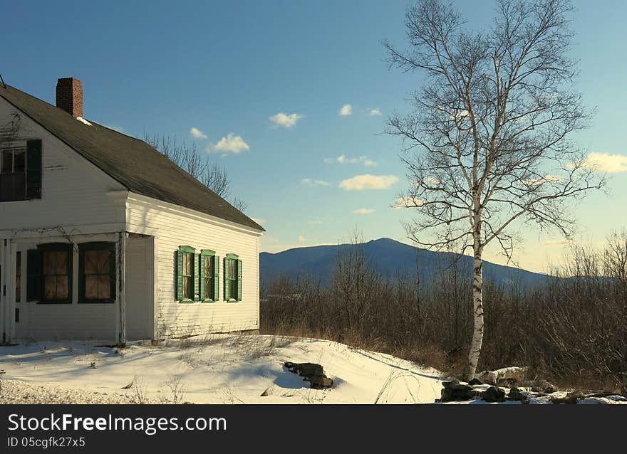 Abandoned house with green shutters in snow with blue sky and birch tree and mountain in background. Abandoned house with green shutters in snow with blue sky and birch tree and mountain in background