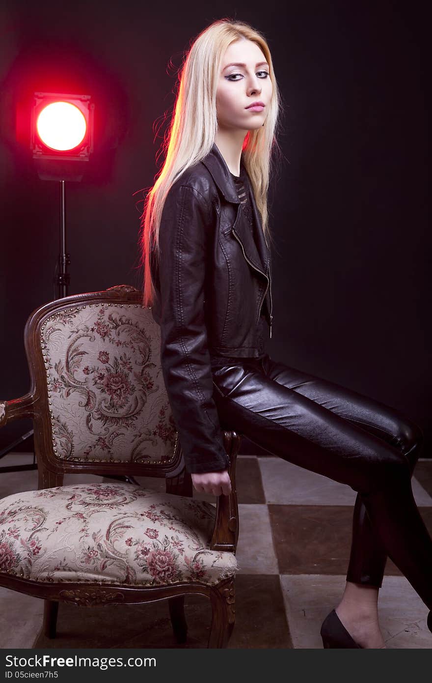 Girl in leather jacket with a red light behind sitting next to a vintage chair studio shot