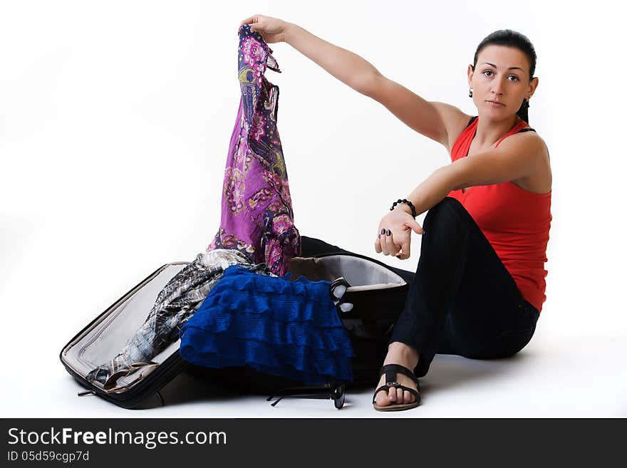 Portrait of young woman sitting next to her open suitcase and preparing for her travel. She is wondering which clothes is better to take. Shot in studio. Isolated over white. Portrait of young woman sitting next to her open suitcase and preparing for her travel. She is wondering which clothes is better to take. Shot in studio. Isolated over white.