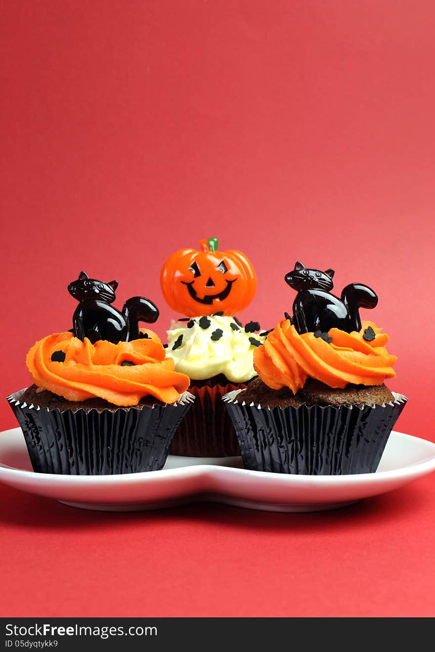 Happy Halloween orange and black decorated cupcakes with black cats and jack-o-lanterns on white heart plate against a red background. Happy Halloween orange and black decorated cupcakes with black cats and jack-o-lanterns on white heart plate against a red background.