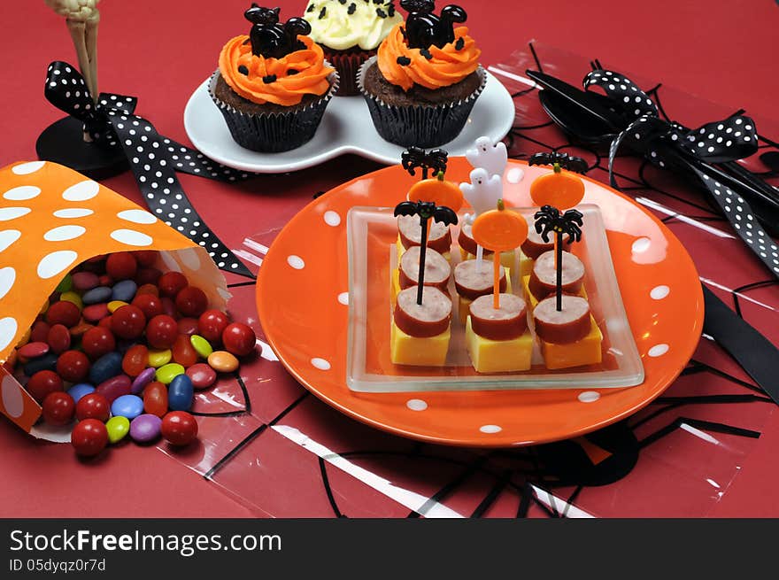 Happy Halloween party table with skeleton glass, cupcakes, candy lollies and party food with orange and black pumpkin, cat, bat and ghost decorations.