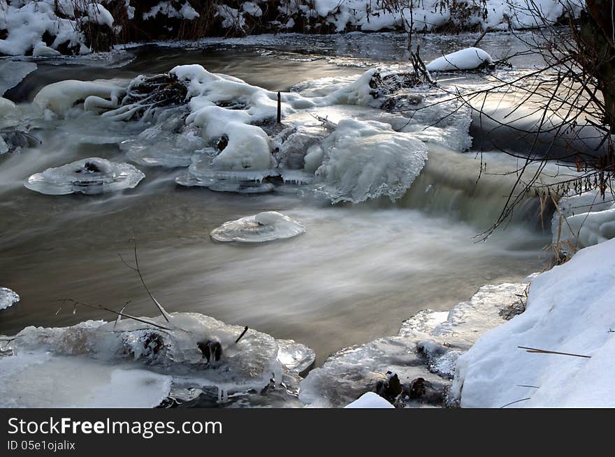 The photograph shows a small waterfall on the river in winter scenery. The banks of the river are covered with a layer of snow. In the water we see the ice caps. The photograph shows a small waterfall on the river in winter scenery. The banks of the river are covered with a layer of snow. In the water we see the ice caps.