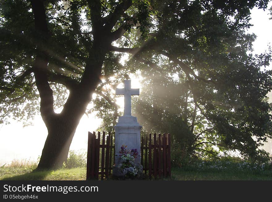 The photograph shows a roadside old concrete cross, standing in the shade of a tree. It's morning, hovering above the ground fog lightened glow of the rising sun. The photograph shows a roadside old concrete cross, standing in the shade of a tree. It's morning, hovering above the ground fog lightened glow of the rising sun.