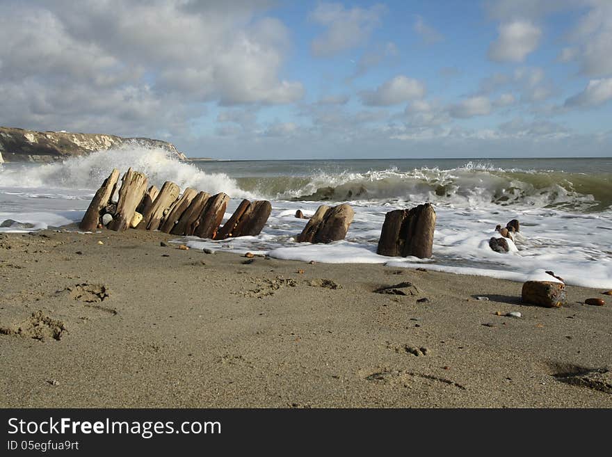 Waves crashing onto the remains of an old sea defence on a sandy beach. Cliffs and sky in the background. Waves crashing onto the remains of an old sea defence on a sandy beach. Cliffs and sky in the background