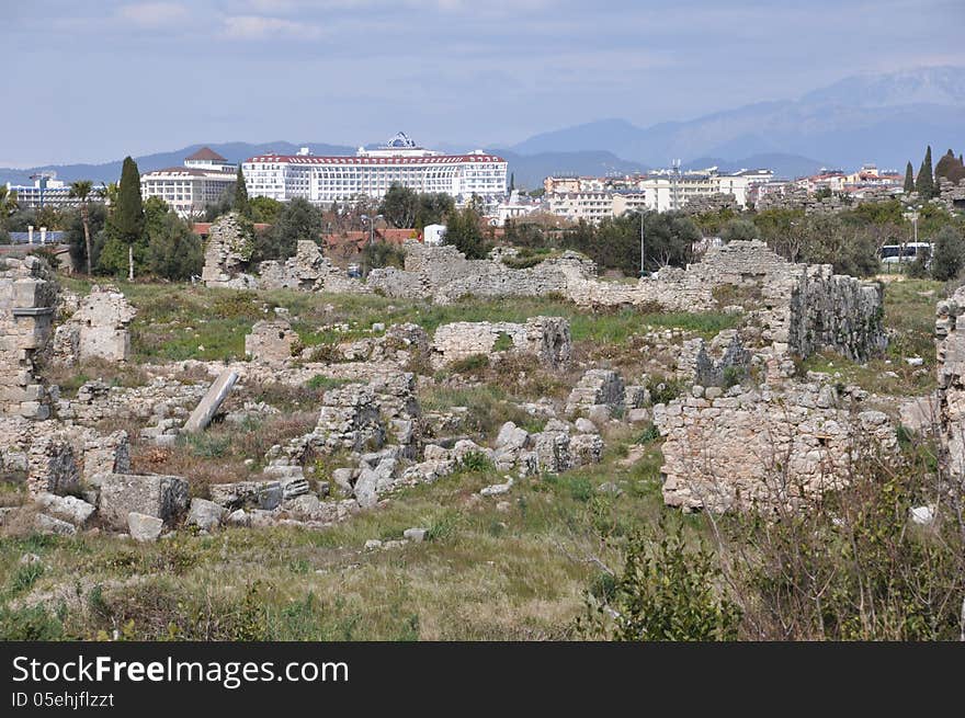 Image shows details of Antique Side, Turkey, with some hotels in the back. Image is dominated by ruins in the front and in the middle. Sky is blue and light cloudy. Image shows details of Antique Side, Turkey, with some hotels in the back. Image is dominated by ruins in the front and in the middle. Sky is blue and light cloudy.