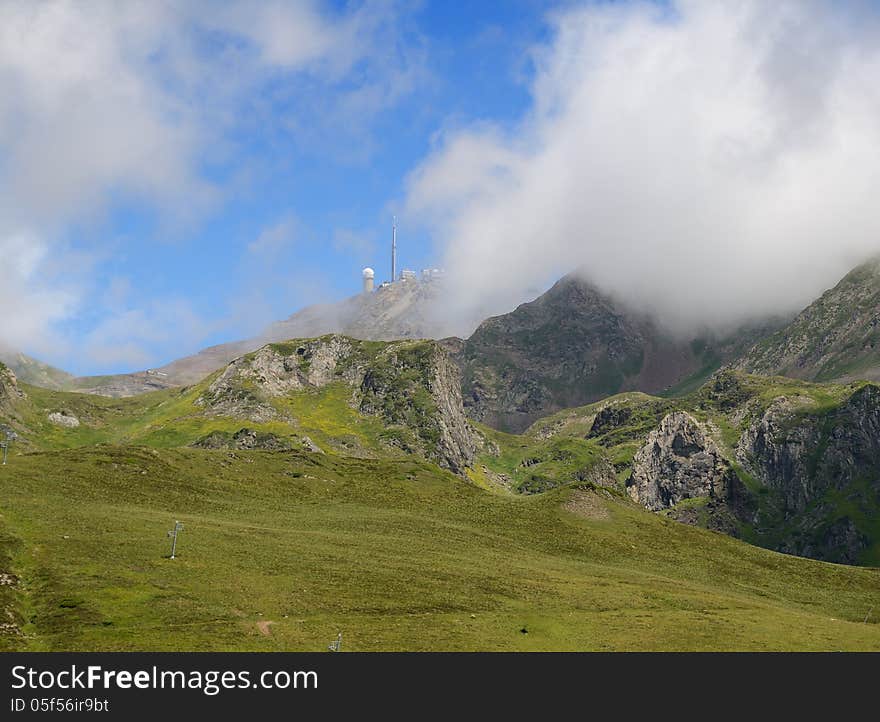 There are green slopes of mountains from the pass of Tourmalet in Pyrenees. In the background the remote peak (Pic du Midi) with an astronomical observatory is covered with white clouds. There are green slopes of mountains from the pass of Tourmalet in Pyrenees. In the background the remote peak (Pic du Midi) with an astronomical observatory is covered with white clouds.