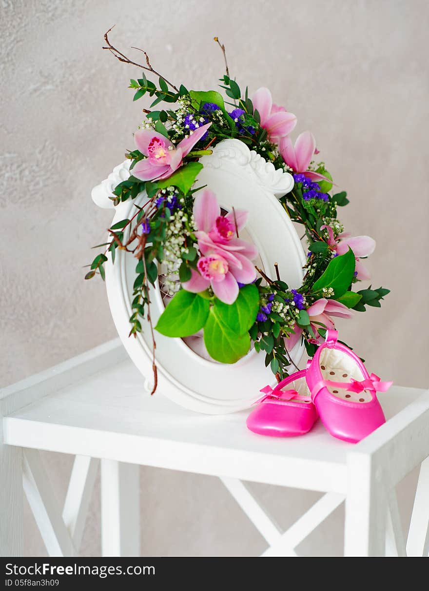 Still life with orchid wreath hanging on the retro style photo frame and infant shoes standing on white wooden stand. Still life with orchid wreath hanging on the retro style photo frame and infant shoes standing on white wooden stand