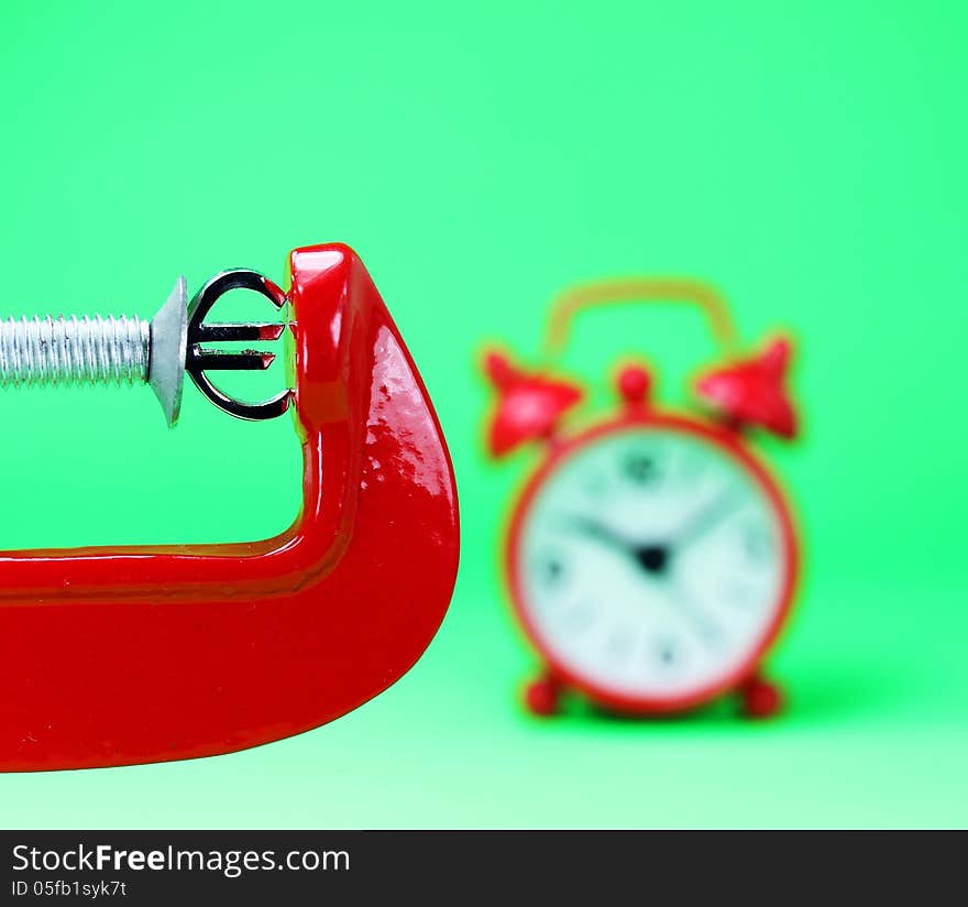 A silver Euro symbol placed in a red clamp with a pastel green background, with a red alarm clock in the background indicating the pressure on the Euro. A silver Euro symbol placed in a red clamp with a pastel green background, with a red alarm clock in the background indicating the pressure on the Euro.