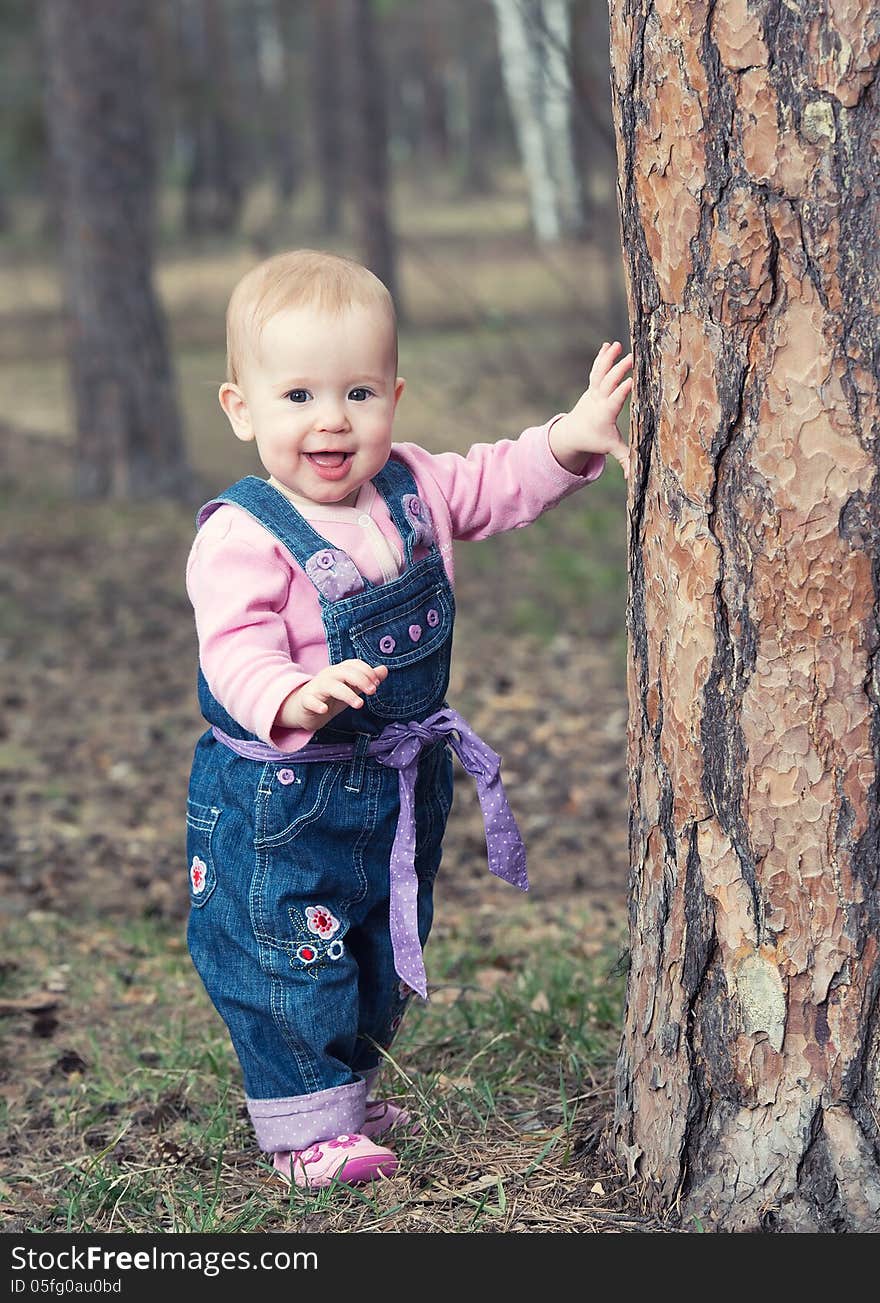 Happy baby girl in jeans jumpsuit stands on legs near a tree in the park outdoors. Happy baby girl in jeans jumpsuit stands on legs near a tree in the park outdoors