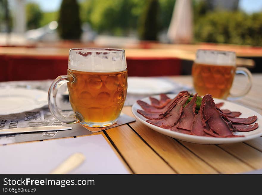 On a table with a white tablecloth is a plate with Basturma and two mugs of beer