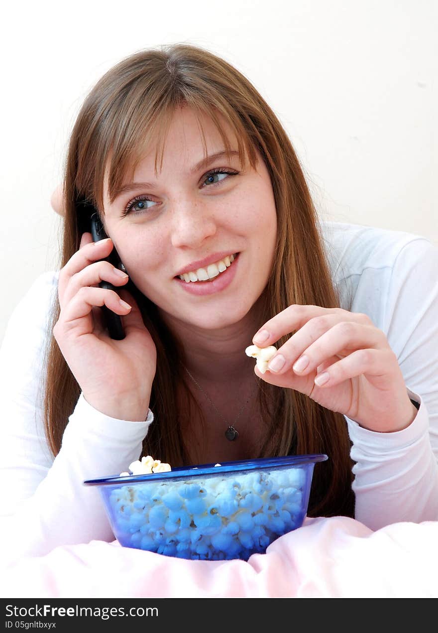 Beautiful young woman enjoying eating popcorn and talking on cell phone on her bed. - Stock Image. Beautiful young woman enjoying eating popcorn and talking on cell phone on her bed. - Stock Image