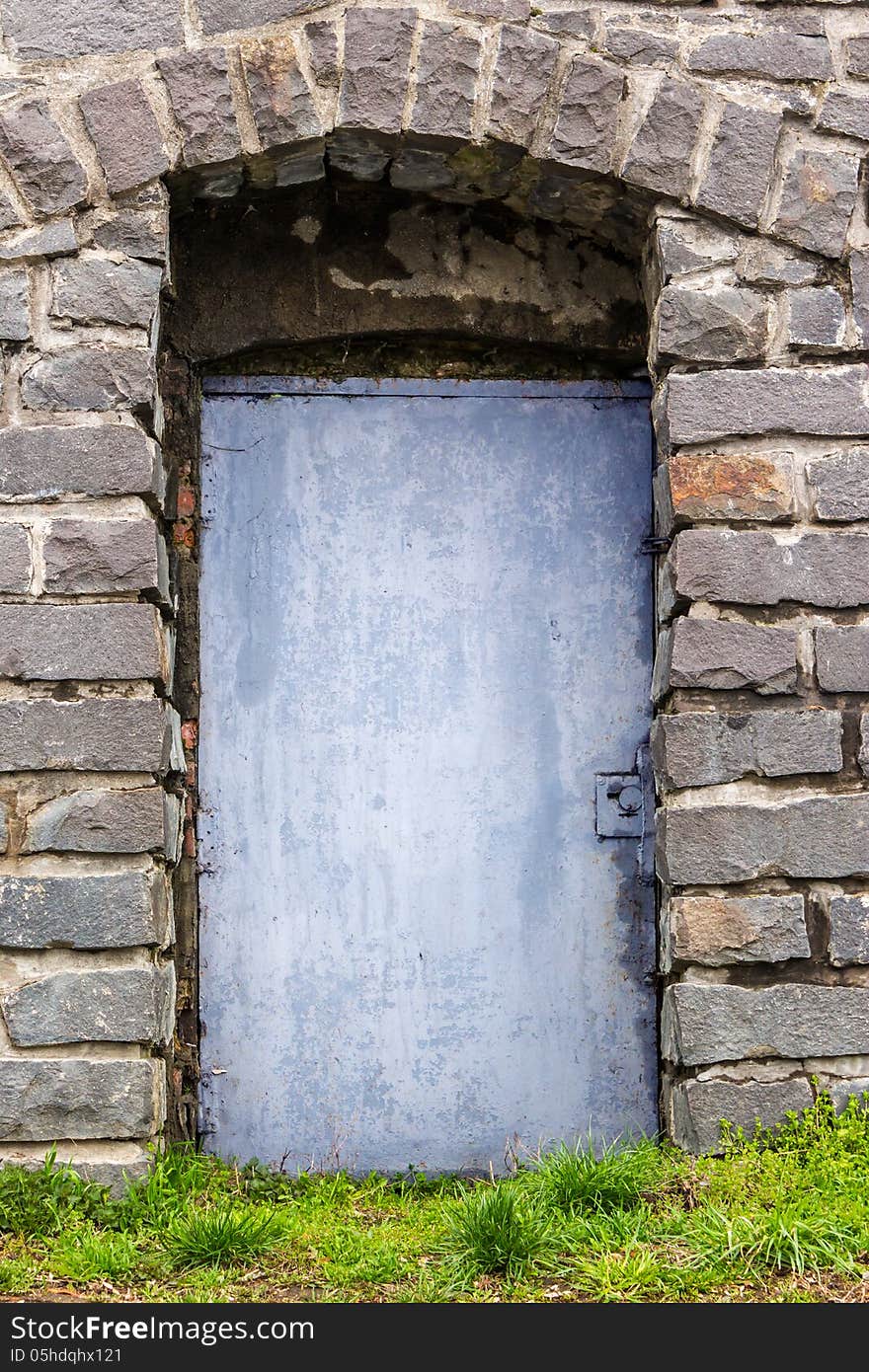 Stone wall with an arch and blue metal door, and the grass in front of it. Stone wall with an arch and blue metal door, and the grass in front of it