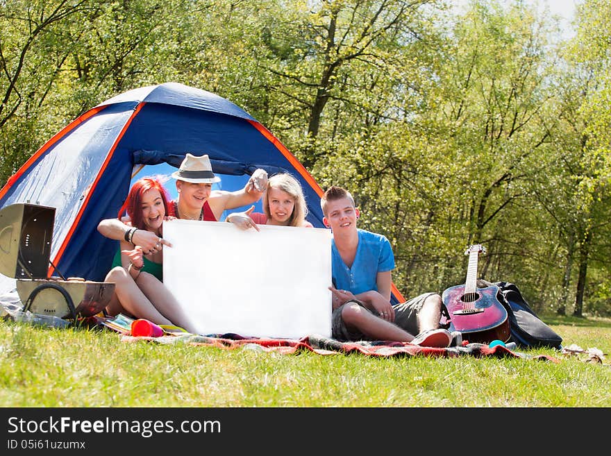 Four teenagers in colored shirts on the camping with a white board which can be used for your advertisements. Four teenagers in colored shirts on the camping with a white board which can be used for your advertisements