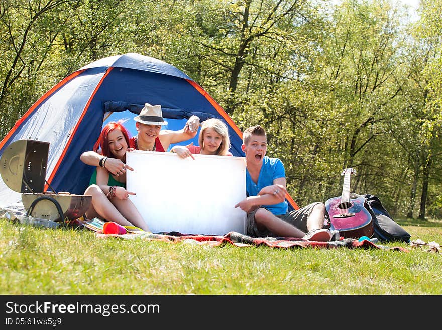 Four teenagers in colored shirts on the camping with a white board which can be used for your advertisements. Four teenagers in colored shirts on the camping with a white board which can be used for your advertisements