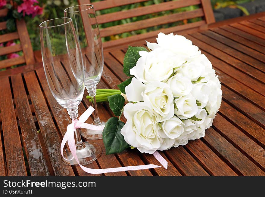 Wedding bridal bouquet of white roses with two champagne glasses with pink polka dot ribbon on outdoor garden table setting after rain.