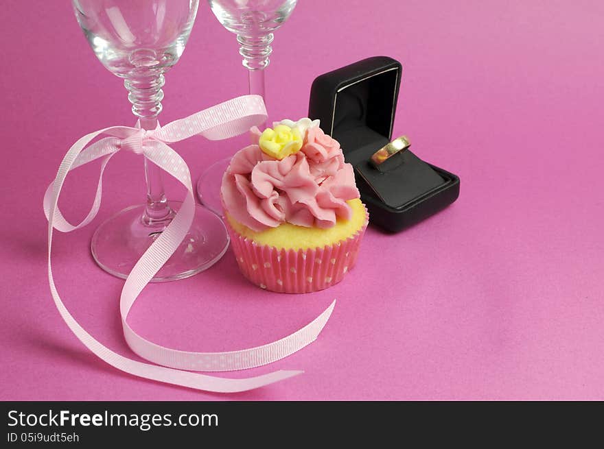 Wedding theme bridal pair of champagne flute glasses with pink cupcake and wedding ring in black jewlery box against a pink background.