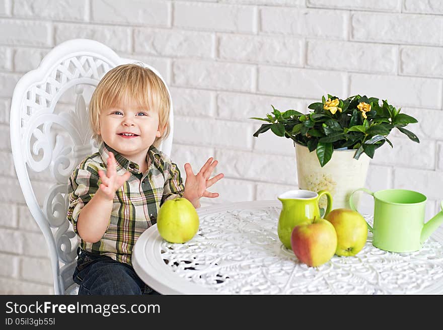 Cheerful baby boy sitting at the vintage style table with pot flower, apples, watering can and a jar. Cheerful baby boy sitting at the vintage style table with pot flower, apples, watering can and a jar