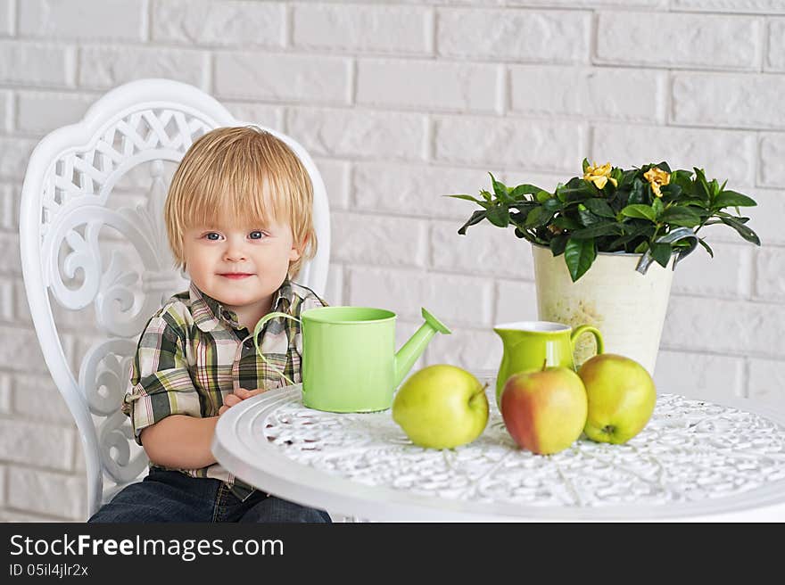 Cute little boy sitting at the vintage style table with pot flower, watering can and apples. Cute little boy sitting at the vintage style table with pot flower, watering can and apples