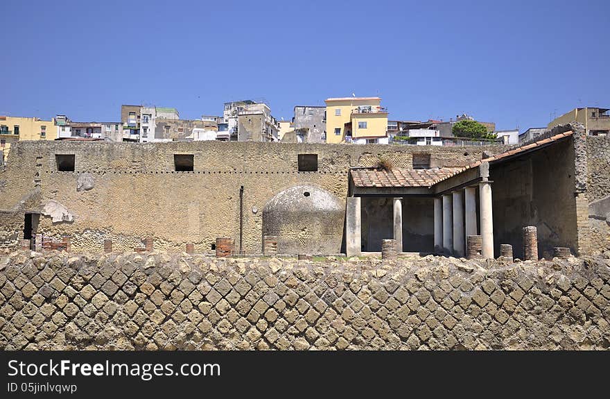 Herculaneum, an ancient Roman town destroyed by volcanic pyroclastic flows from Mount Vesuvius, with modern Ercolano seen against a blue sky behind the bath house. Herculaneum, an ancient Roman town destroyed by volcanic pyroclastic flows from Mount Vesuvius, with modern Ercolano seen against a blue sky behind the bath house.