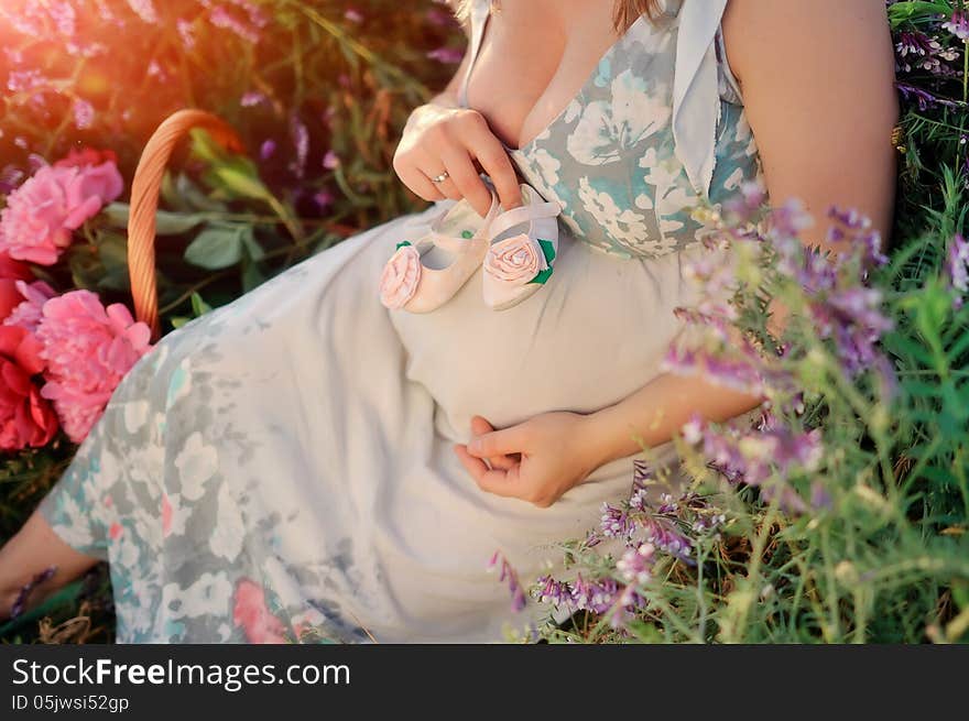 Against the background of a flowering field belly of a pregnant woman with baby shoes in hand. Against the background of a flowering field belly of a pregnant woman with baby shoes in hand