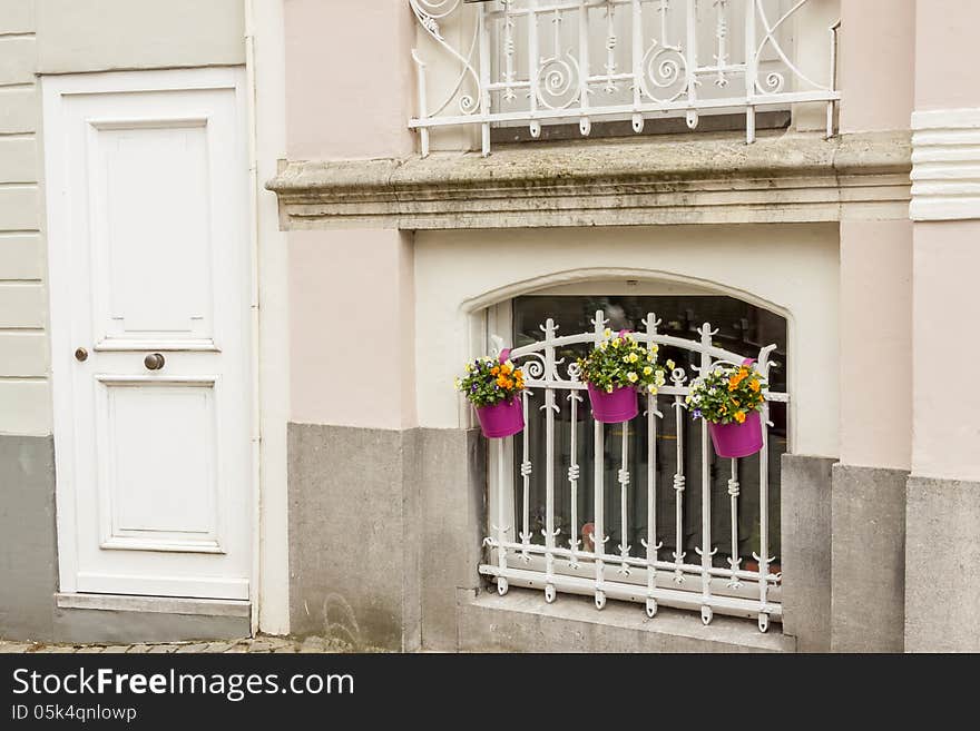 White wooden door and window with bars and three plants in pink flowerpots. Brugge old Town - Belgium. White wooden door and window with bars and three plants in pink flowerpots. Brugge old Town - Belgium.