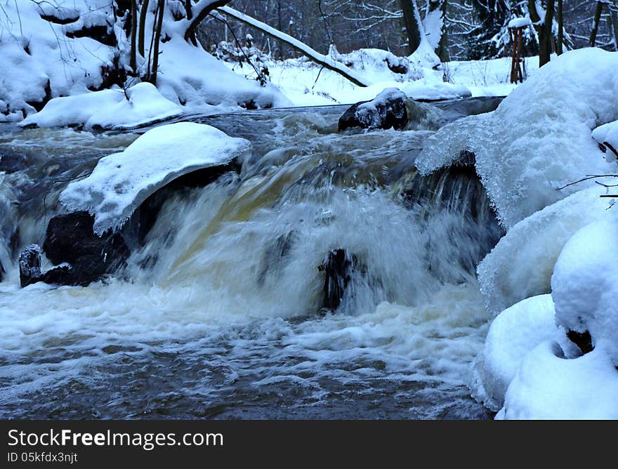 The photograph shows a small waterfall. It is winter on the banks and in the surrounding trees behind a layer of snow. The photograph shows a small waterfall. It is winter on the banks and in the surrounding trees behind a layer of snow.