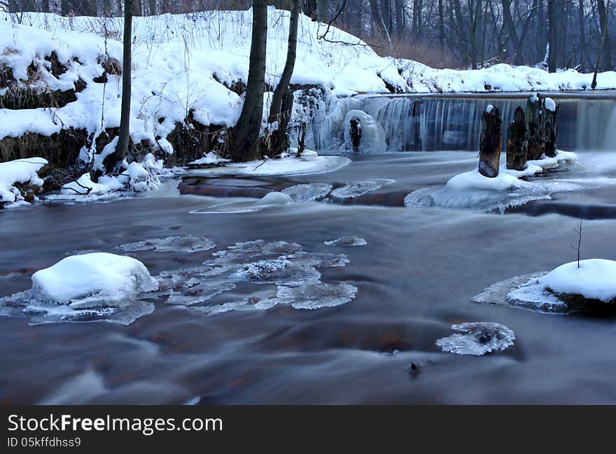 The photograph shows a small waterfall. It is winter on the banks and in the surrounding trees behind a layer of snow. The photograph shows a small waterfall. It is winter on the banks and in the surrounding trees behind a layer of snow.