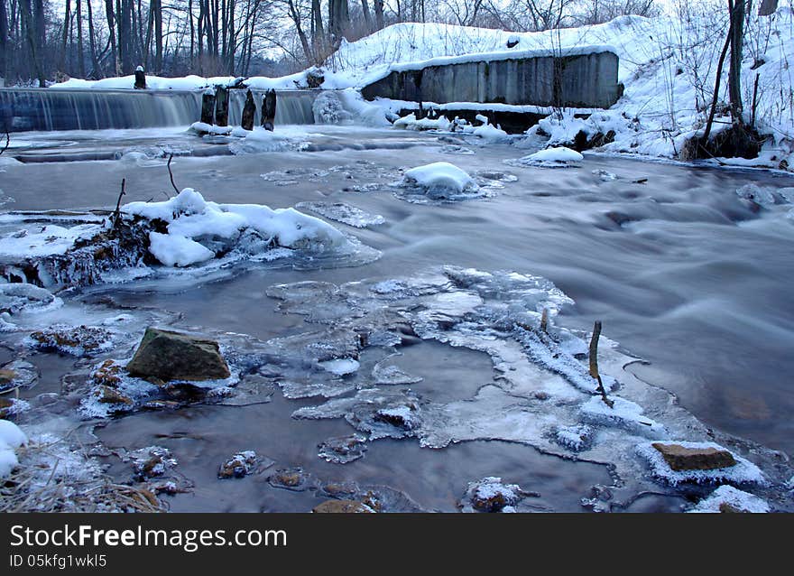 The photograph shows a small waterfall. It is winter on the banks and in the surrounding trees behind a layer of snow. The photograph shows a small waterfall. It is winter on the banks and in the surrounding trees behind a layer of snow.