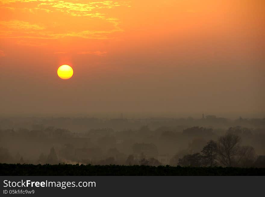 The photograph shows the evening sky, the sun standing low on the horizon. In the valley we see the roofs of houses out there that are steeped in the evening mist. The photograph shows the evening sky, the sun standing low on the horizon. In the valley we see the roofs of houses out there that are steeped in the evening mist.