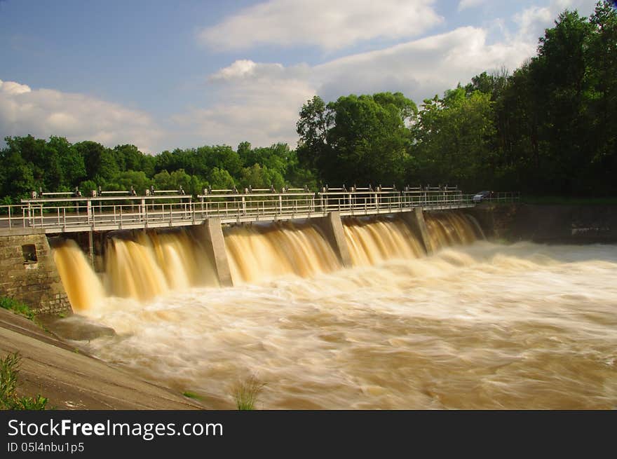 The photograph shows a dam on the river during high water levels. It takes dump excess water flows over the dam increased amount of brownish white water. The photograph shows a dam on the river during high water levels. It takes dump excess water flows over the dam increased amount of brownish white water.