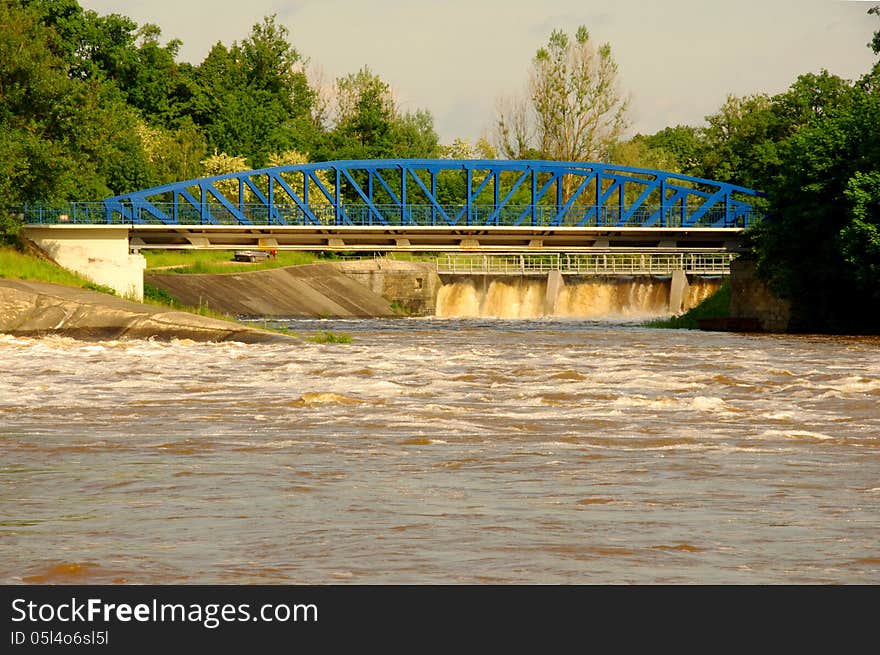 The photograph shows a dam on the river during high water levels. It takes dump excess water flows over the dam increased amount of brownish white water. The photograph shows a dam on the river during high water levels. It takes dump excess water flows over the dam increased amount of brownish white water.